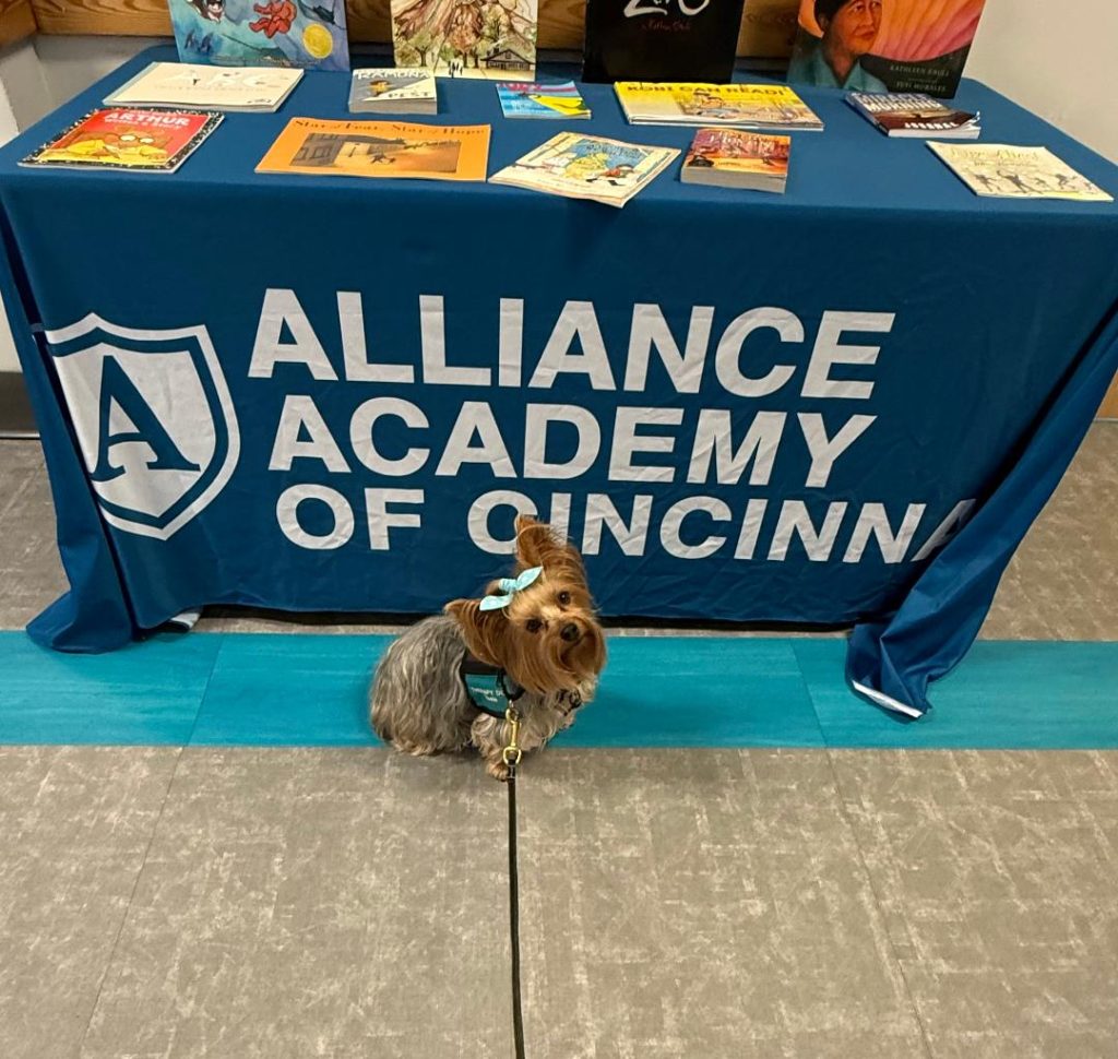 A small dog with a blue bow sits on the floor in front of a table covered with a blue Alliance Academy of Cincinnati tablecloth. Books are displayed on top of the table.