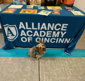 A small dog with a blue bow sits on the floor in front of a table covered with a blue Alliance Academy of Cincinnati tablecloth. Books are displayed on top of the table.