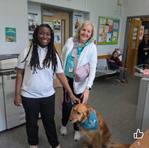 Two women stand smiling next to a golden retriever wearing a blue bandana in a community center or office. In the background, people sit and bulletin boards are visible on the wall.