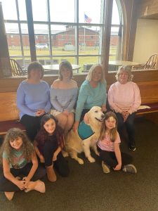 Six women and girls sit and kneel around a golden retriever wearing a teal vest inside a room with large windows. An American flag is visible outside in the background. Everyone is smiling.