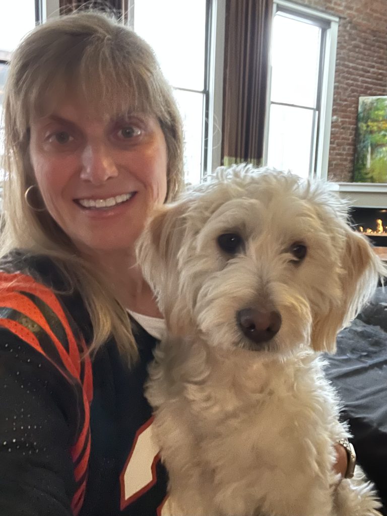 A woman with long blonde hair smiles at the camera while holding a fluffy white dog. They are indoors, with large windows, brown curtains, and a brick wall with a fireplace in the background.