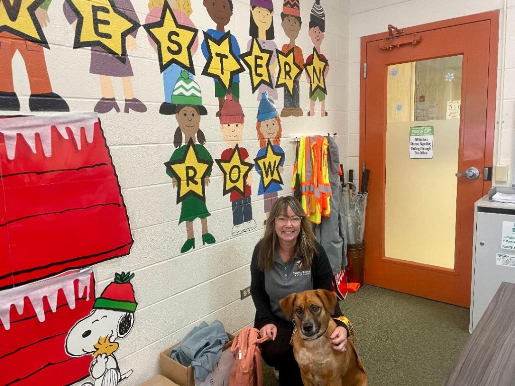 A woman sits indoors next to a brown dog, smiling, in front of a colorful wall mural with cartoon children and stars spelling Western Row. Reflective vests and jackets are hanging nearby.