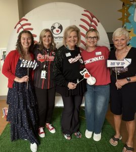 Five women stand smiling in front of a large baseball backdrop. They wear sports-themed clothing, mostly in red and black, and hold props like “GO TEAM,” “Cincinnati,” and “MVP.” The setting has a fun, celebratory atmosphere.