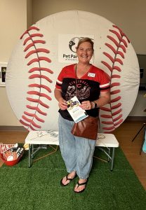 A smiling woman stands in front of a large baseball backdrop, holding brochures. She wears a Cincinnati Reds shirt, jeans, and a crossbody bag. A bench, baseball-themed props, and a Pet Partners sign are visible.