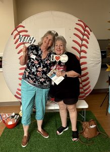 Two smiling women stand on fake grass in front of a large baseball backdrop, holding sports-themed props, including Go Team and I Love Home. A brown purse sits on the floor beside them.