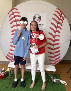 A teenage boy and a woman stand smiling in front of a large baseball backdrop, holding props and a trophy. The boy wears a blue hoodie and shorts; the woman wears a red shirt and white pants. Both have name tags.