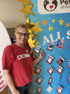 A smiling woman in a red Cincinnati shirt stands next to a decorated display board with gold stars, pet photos, and ID cards at a Pet Partners event.