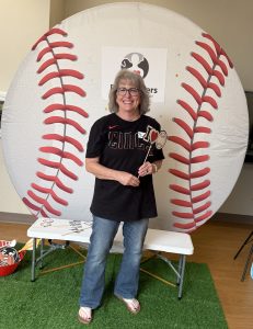A smiling woman stands in front of a large baseball-themed backdrop, holding photo props. She wears glasses, a black t-shirt, jeans, and sandals. There is a bench with props and a grass-like mat beneath her.