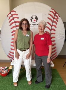 Two women stand smiling in front of a large baseball backdrop with the Pet Partners Greater Cincinnati logo. One wears a green top and white pants, the other a red shirt and gray pants. They both wear name tags.