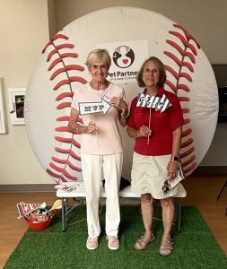 Two women stand in front of a large baseball backdrop. One holds a MVP sign, the other holds a GO TEAM sign. They are smiling, and theres a basket of props on the left and a Pet Partners logo above them.