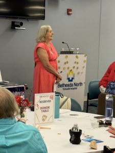 A woman in a coral dress stands and speaks at a podium in a hospital event room. The podium has a Bethesda North TriHealth Hospital sign. A table in the foreground has a Reserved Donor Table sign and several decorations.