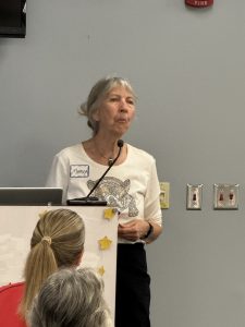 An older woman with gray hair speaks at a podium, wearing a name tag and a shirt with an animal graphic. A microphone is clipped to her collar, and people are seated in front of her. The background is a plain wall with light switches.