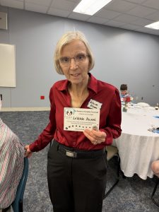 An older woman with short gray hair, glasses, and a red blouse stands indoors, holding a name tag that reads LaVerne Allan. She is near a round table with decorations and people seated in the background.