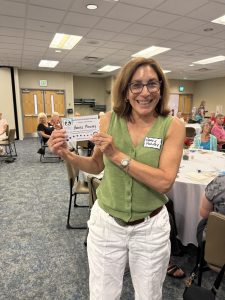 A smiling woman with glasses, wearing a green vest and white pants, holds up a certificate. She stands in a conference room with a group of people seated at round tables in the background.