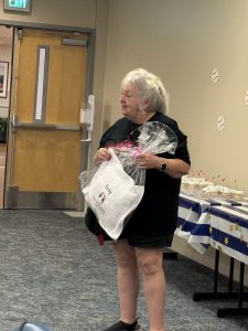 An older woman with short gray hair stands indoors holding a white tote bag and a wrapped gift. She wears a black shirt and shorts. Behind her is a table with cupcakes and a blue star-patterned tablecloth.