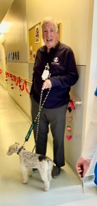 An older man smiles while holding a small dog on a leash in a hallway decorated with red garlands and paper hearts. He wears a dark sweatshirt with an ID badge. Part of another person is visible on the right.