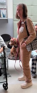 A woman in tan clothes stands in a hospital room holding a leash attached to a small dog with red ribbons on its ears, sitting on a chair. A person in a hospital gown and another person are partially visible nearby.
