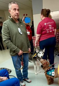 A man stands indoors holding a leash attached to a Husky therapy dog. Several people, one in a red shirt with logos and one with a blue face, stand nearby. Another small dog wearing a vest is in the foreground.