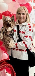 A smiling woman wearing a white jacket with red hearts holds a small dog. Red and pink balloons are in the background, along with a large white sign featuring handwritten messages.