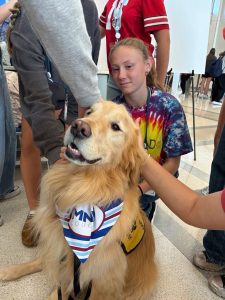 A golden retriever wearing a striped bandana sits as several people pet it. A girl in a tie-dye shirt is kneeling beside the dog, smiling. The scene appears to be indoors, possibly at a school event or fair.