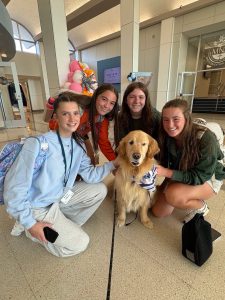 Four smiling young women crouch indoors, petting a golden retriever wearing a shirt and lanyard. The group appears happy, surrounded by bright decor and natural light in a spacious, modern lobby.
