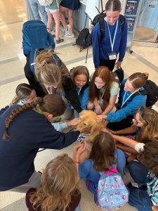 A group of smiling girls kneels in a circle, petting a golden retriever indoors. Some wear blue shirts and lanyards, and backpacks are visible. The mood appears joyful and friendly.
