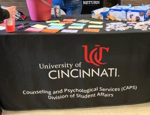 A table covered with a University of Cincinnati Counseling and Psychological Services (CAPS) cloth, featuring colorful sticky notes, markers, stickers, snacks, and a pink bucket.